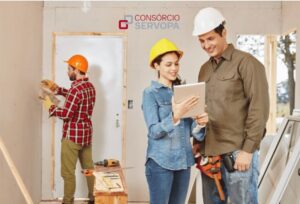 Arquiteta e mestre de obras olhando o projeto em uma sala em reforma, ao fundo homem de luvas olhando na parede onde vai fazer um furo. com a logo do consorcio servopa acima de todos.